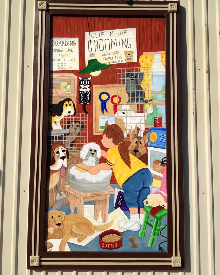 Painted outdoor mural of a groomer bathing a poodle in a tub, with boarding and grooming signs, on the shop's metal wall